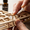 The person is applying glue to a small wooden piece. The finished parts of the model have a clean, natural, and seamless look, with no glossy or shiny glue residue visible