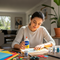 A bright, airy lifestyle image featuring a young woman smiling as she works on a creative DIY project at a wooden table. She is holding a bottle of Glues Australia PVA glue, surrounded by colorful craft supplies, to show the product